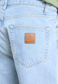 Jeans in denim azzurro chiaro con tasca a toppa. Caratteristiche notevoli includono una patch con logo in pelle marrone e dettagli in cucitura gialla.