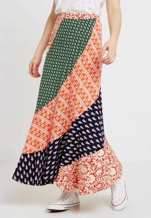 Kvinde iført en flerfarvet patchwork maxi nederdel med grønne, orange, marineblå og cremefarvede blomsterprint, kombineret med hvide sneakers.