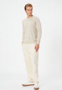 Helles beige Strickpullover mit strukturiertem diagonalen Muster und gerippten Bündchen, kombiniert mit weit geschnittenen cremefarbenen Hosen und weißen Sneakers.