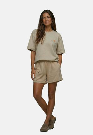 Femme debout les mains dans les poches, portant une chemise beige à manches courtes, un short assorti et des chaussures marron sans lacets sur un fond uni.