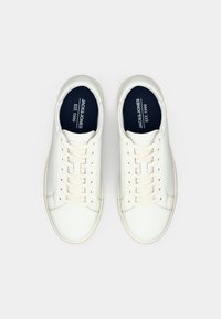 Par bijelih kožnih tenisica na vezanje s natpisom "JACK & JONES EST. 1990" otisnutim na crnim ulošcima, prikazano odozgo na bijeloj pozadini.