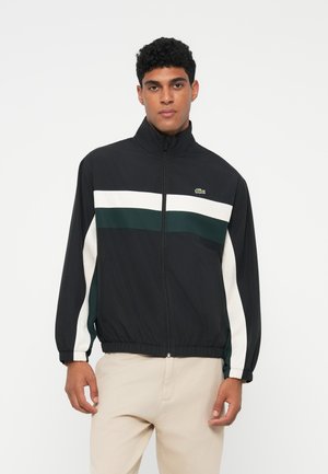 Giacca nera con strisce bianche e verdi, chiusura a zip, colletto rialzato, polsini e orlo elasticizzati, con un piccolo logo Lacoste sul petto.