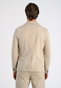 Giacca beige su misura realizzata in tessuto texturizzato, con colletto a punta, spacco posteriore singolo e dettagli neri sui bottoni delle maniche.