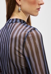 Top trasparente con strisce verticali in tonalità di marrone e blu. Tessuto leggero e testurizzato con scollatura alta e maniche lunghe. Orecchini pendenti in argento.