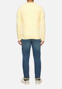 Hellgelber gestrickter Pullover mit langen Ärmeln, Rundhalsausschnitt und gerippten Säumen, kombiniert mit blauen Jeans und weißen Sneakers.