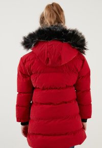 Rød pufferjakke med sort faux fur kant på hætten, kviltet design og elastiske manchetter. Materialet fremstår skinnende og isoleret.