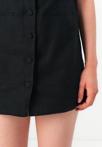 Robe en lin noir avec un devant à boutons, de longueur courte et deux poches latérales. Le tissus présente une texture subtile et des détails de couture minimaux.