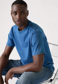 Blå bomull T-skjorte med korte ermer, rund hals og subtil logo detalj. Brukt med denim jeans mens man sitter på en klar stol.