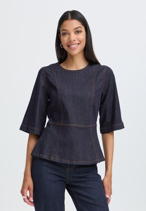 Femme aux longs cheveux foncés souriante portant un haut péplum en denim foncé avec des manches longueur coude et un jean en denim foncé assorti.