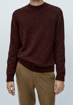 Person, die einen dunkelroten, gesprenkelten Strickpullover und hellbraune Hosen trägt, steht mit leicht angewinkelten Armen seitlich vor einem schlichten Hintergrund.