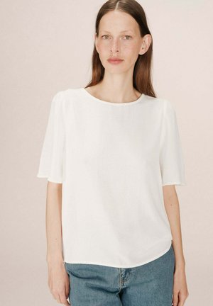 Blouse blanche à manches courtes avec un col rond, texture douce et manches légèrement bouffantes, associée à un jean en denim clair.