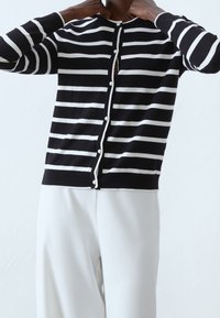 Černobílý pruhovaný kardigan s kulatým výstřihem, zapínáním na knoflíky a perličkovými knoflíky. Měkký pletený materiál s hladkou texturou.