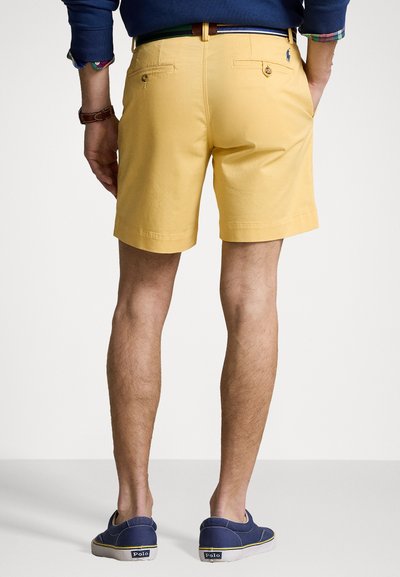 Bermudas amarelas em algodão com bolsos traseiros e detalhe em botão, combinadas com sapatos slip-on azul marinho. As bermudas têm uma bainha dobrada e têm um comprimento até a metade da coxa.