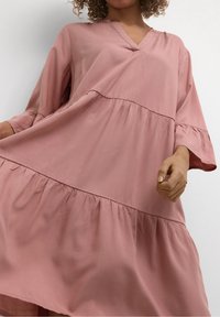 Pitkä vaaleanpunainen mekko pehmeästä kankaasta, jossa on V-kaula-aukko, pitkät hihat ja kerroksellinen rypytyshelma. Sileä pinta ja rento istuvuus.