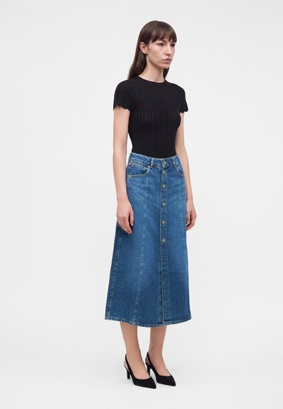 Femme portant un top noir ajusté à manches courtes, une jupe midi en denim bleu à boutons sur le devant et des chaussures slingback noires à talons bas, debout de profil.
