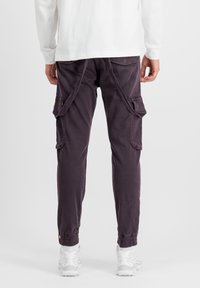 Cargo-Hosen aus einem dunkelvioletten Baumwollgemisch, mit seitlichen Taschen, schmal zulaufendem Bein, elastischen Bündchen und einem lockeren Schnitt. Getragen mit weißen Sneakern.