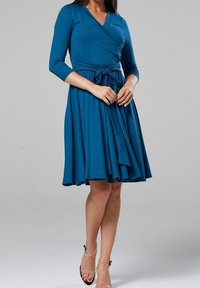 Teal omlottklänning med trekvartsärmar, bälte i midjan och utsvängd kjol. Släta tyget med ett flytande mönster och knälång fåll.