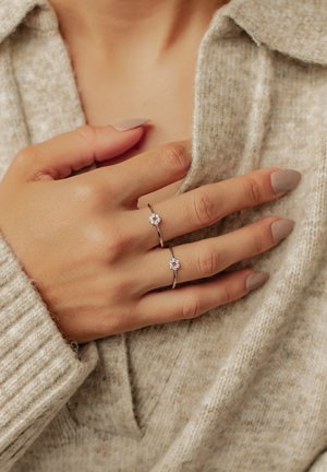 Two silver rings with round, clustered clear gemstones on a hand, set against a textured beige sweater. Nails are light beige.