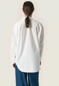 Hvid skjorte med lange ærmer, afslappet pasform, buet kant og blød tekstur, parret med wide-leg blå denimbukser.