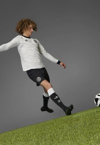 White long-sleeve jersey with black accents, black shorts, and knee-high black socks with white bands; kicking a soccer ball on grassy surface.
