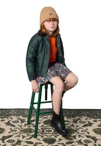 Veste matelassée verte, sweat à capuche graphique orange, jupe florale, bottines noires sur un tabouret vert. Bonnet en tricot beige. Tapis texturé en dessous.