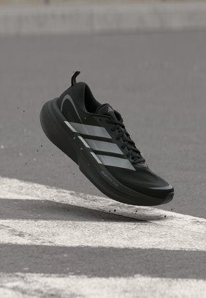 Chaussure de course Adidas noire avec des bandes argentées et le texte « Dreamstrike Glow », suspendue en l'air au-dessus de l'asphalte et des marquages blancs sur la route.