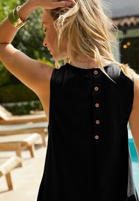 Haut sans manches noir avec un col rond et cinq boutons en bois dans le dos, offrant une coupe décontractée et de légentes fentes sur les côtés.