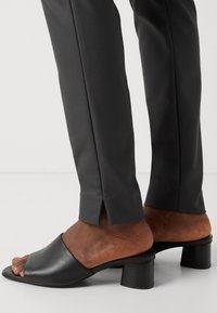 Schwarze Leder-Sandalen mit offenem Zehenbereich und niedrigem Blockabsatz. Kombiniert mit schmal geschnittenen grauen Hosen, die einen dezenten Schlitz am Saum haben.