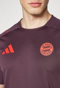 FC Bayern München tröja i mörklila med orange logotyp och tre-randig design på axeln. Tillverkad av tunt, stretchigt material.