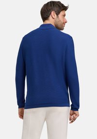 Niebieski sweter dzianinowy o teksturowanym wykończeniu, z kołnierzykiem oraz ściągaczami na rękawach i dolnej krawędzi, noszony na jasnych spodniach.