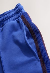 Kék sportnadrág elasztikus derékpánttal, sötétlila texturált hálós oldalsávval és oldalzsebbel.