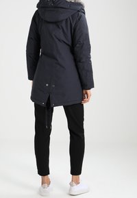 Granatowa puchowa kurtka z kapturem, ozdobiona futerkiem z imitacji, z taliowanym krojem, bocznymi kieszeniami oraz rozcięciem z tyłu. Noszona z czarnymi spodniami.