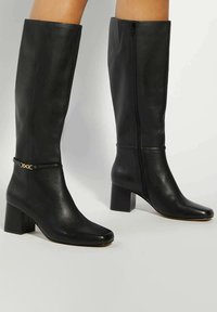 Black leather knee-high boots with a square toe, block heel, side zipper, and a decorative gold chain accent near the ankle.