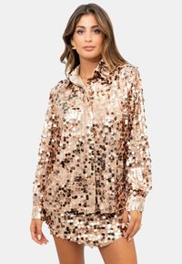 Top in lamè rosa con paillettes, con colletto, maniche lunghe e vestibilità rilassata. Presenta un mix di paillettes grandi e piccole per dare texture.