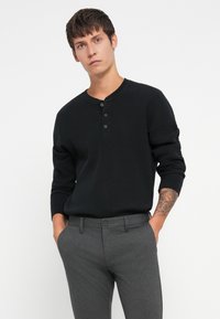 Camisa henley de manga larga negra con cuello de botones, con tres botones, combinada con pantalones grises. Enfocarse en la textura y el diseño ajustado.