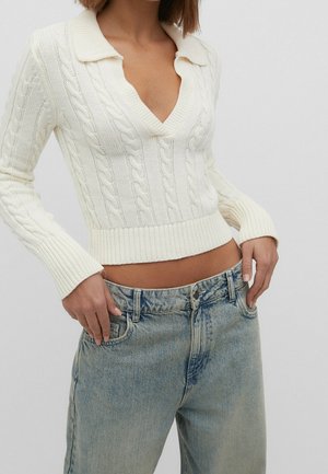Femme portant un pull en maille torsadée blanc avec un col en V profond et un jean taille haute bleu clair, mains dans les poches avant, sur un fond uni.