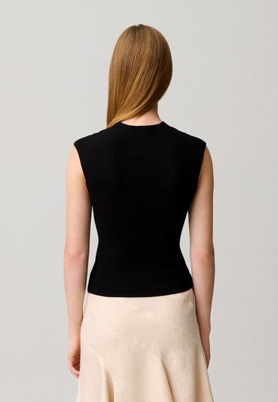 Donna con capelli lisci castano chiaro indossa top nero senza maniche e gonna beige chiaro, vista di spalle su sfondo neutro.