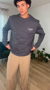 Noor mees lokkis juustega, kandes tumehall Allsaintsi pusa ja beeže pükse, seisab valgusküllases toas taimede keskel.