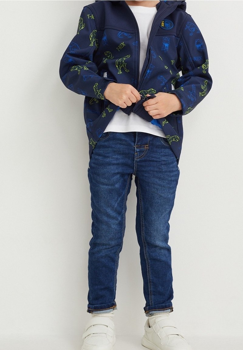Enfant portant une veste bleue à motif dinosaure, un t-shirt blanc, un jean bleu et des baskets blanches, en train de fermer la fermeture éclair de la veste.