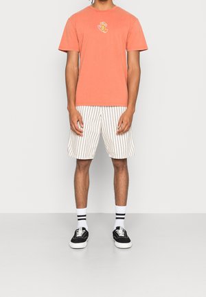 Koralowy t-shirt z krótkim rękawem z nadrukiem na piersi, w połączeniu z białymi szortami w paski, czarnymi trampkami i sportowymi skarpetami.