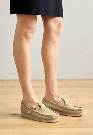 Beige suede shoes with a rounded shape, featuring a T-strap and buckle, resting on a soft sole, viewed from the legs down.