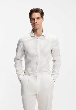 Man with dark hair wearing a white and beige vertical striped button-up shirt and white trousers, hands in pockets, against plain white background.