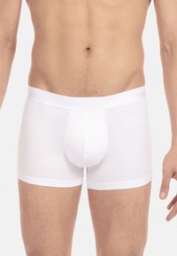 Männlicher Oberkörper, der schlichte, weiße, enganliegende Boxer-Briefs trägt, steht vor einem neutralen, hellen Hintergrund mit entspannten Armen an den Seiten.