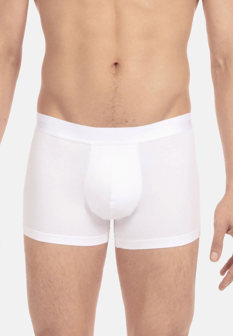 Männlicher Oberkörper, der schlichte, weiße, enganliegende Boxer-Briefs trägt, steht vor einem neutralen, hellen Hintergrund mit entspannten Armen an den Seiten.