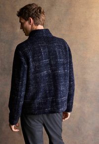 Mann in einer dunkelblauen strukturierten Jacke und grauen Hosen, der steht und mit dem Rücken zu einem neutralen braunen Hintergrund schaut.