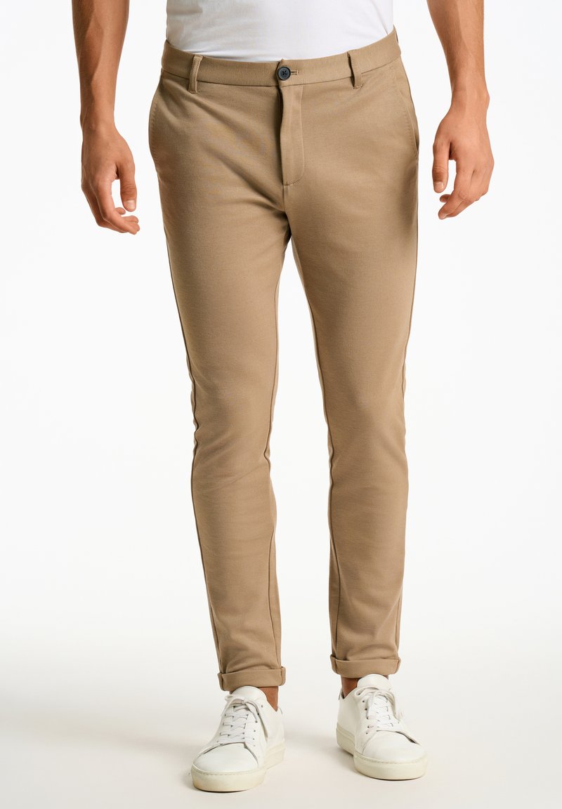 Pantalon slim-fit beige en tissu extensible, avec une fermeture à bouton et des ourlets retroussés, associé à des baskets blanches.