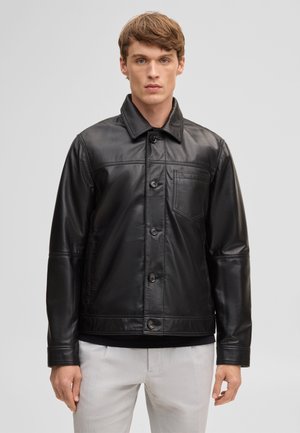 Jeune homme debout face à l'objectif, portant une veste en cuir noire boutonnée avec une poche poitrine et un pantalon gris clair, sur fond uni.