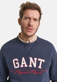 Man met kort bruin haar en lichte stoppelbaard, gekleed in een marineblauwe sweatshirt met het "GANT" logo en een collared shirt eronder, neutrale achtergrond.