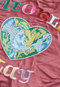 Tessuto trapuntato con finitura in raso rosa che presenta un globo a forma di cuore ricamato multicolore e una tipografia audace e varia.