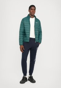 Polstret grønn jakke med høy krage, båret over en hvit skjorte. Navy joggebukser med hvit logo-detalj og svarte joggesko fullfører antrekket.